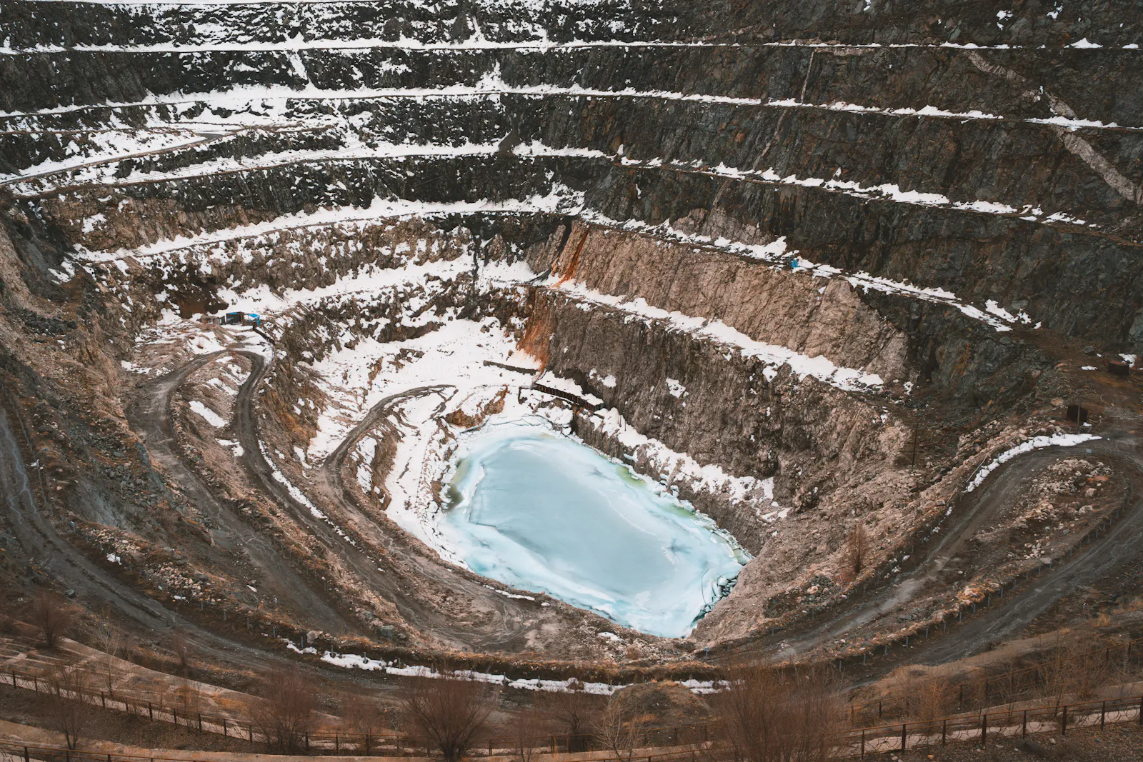 Aerial view of a deep open-pit mine with stepped benches around a pale blue pool